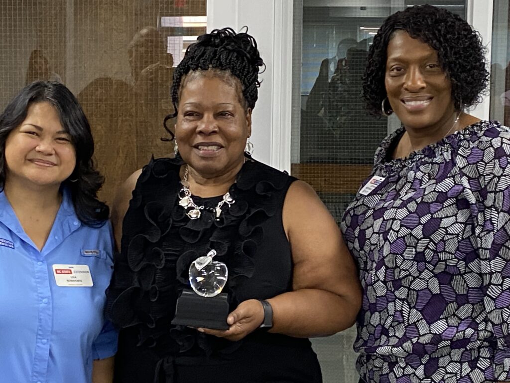 Three women pose together, the middle woman holds a glass trophy.